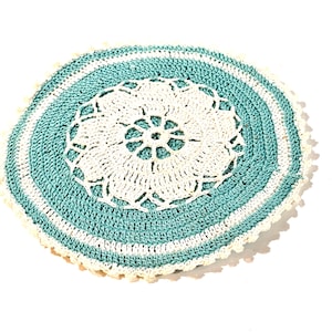 Op de afbeelding: Een rond, met de hand gehaakt kleedje met een centraal wit bloemmotief omringd door concentrische ringen van blauwgroen en wit garen. De buitenrand is afgewerkt met een delicate, geschulpte witte rand, wat een getextureerd, rond textielstuk creëert.