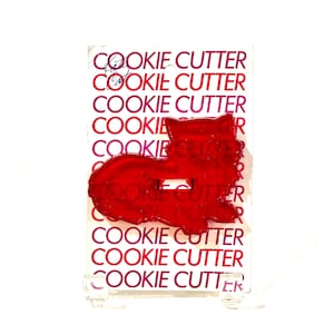 May include: A red plastic cat-shaped cookie cutter attached to a white rectangular backing card. The card features the repeated text "COOKIE CUTTER" in a bold magenta font. The cookie cutter has a small rectangular cutout in the center.