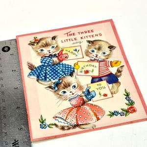 May include: Vintage birthday card featuring three kittens. Each kitten holds a sign with text that reads "Happy Birthday To You". The kittens are wearing dresses and outfits in blue, red, and white. The card has a pink border and floral accents.