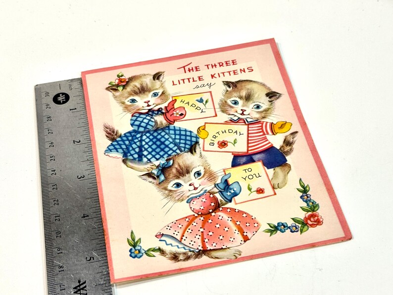 May include: Vintage birthday card featuring three kittens. Each kitten holds a sign with text that reads "Happy Birthday To You". The kittens are wearing dresses and outfits in blue, red, and white. The card has a pink border and floral accents.