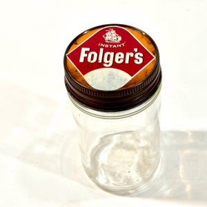 Pode incluir: Um frasco de vidro transparente com tampa de metal. A tampa apresenta um rótulo vermelho e branco com o texto "INSTANT Folger's" e uma ilustração de um navio. O frasco está vazio e parece vintage.