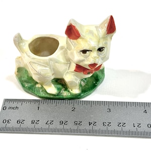 Pode incluir: Estatueta de gato vintage em cerâmica, pintada em branco sujo com detalhes vermelhos nas orelhas e uma gravata borboleta vermelha. O gato está sentado em uma base verde, e mede cerca de 7.6 cm de comprimento.