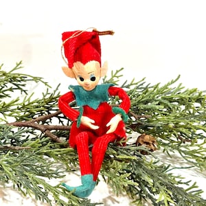 Puede incluir: Adorno navideño vintage de elfo. El elfo viste un atuendo de pana rojo, un cuello y puños verdes, y un sombrero a juego. El elfo tiene una cara pintada y está sentado sobre vegetación. Una pequeña etiqueta está adherida al sombrero.