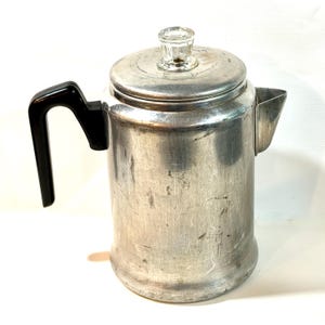 May include: A silver-colored aluminum percolator coffee pot featuring a black plastic handle, a matching aluminum lid, and a clear glass knob on top. The cylindrical body shows signs of wear with visible surface scratches and a metal spout.