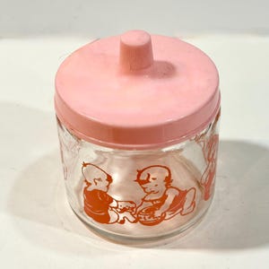 Op de afbeelding: Een vintage glazen pot met een roze deksel en een kleine knop. De pot heeft oranje illustraties van baby's. De pot is helder, waardoor de inhoud zichtbaar is. Een charmant, nostalgisch stuk.