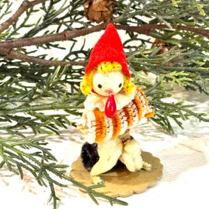 May include: A vintage Christmas ornament featuring a whimsical gnome figure. The gnome has a red felt hat, yellow hair, and a striped orange and brown body. It is mounted on a scalloped cardboard base and set against a backdrop of evergreen branches.