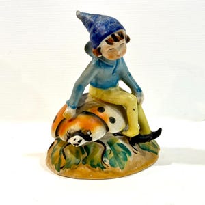 May include: A ceramic figurine of a seated gnome wearing a blue hat and shirt, yellow pants, and black shoes. The gnome is perched on a ladybug with orange and black wings. The base is a tan color with green leaves.