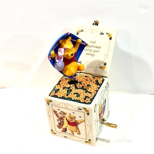 May include: A ceramic music box featuring Winnie the Pooh and Piglet holding a blue umbrella. The lid interior reads, Let happiness carry you away. The exterior displays illustrations of Pooh and Tigger, with a gold-toned metal crank on the side.