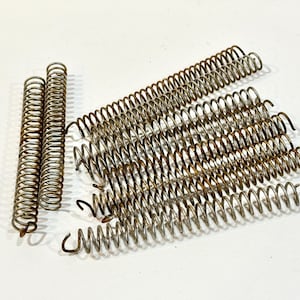 May include: A collection of antique metal springs in various sizes and shapes. The springs are made of a tarnished, aged metal, with a spiral design. Some are tightly coiled, while others are more elongated. The springs are arranged on a white surface.