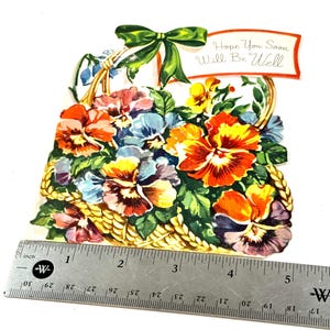 May include: A vintage greeting card featuring a basket overflowing with colorful pansies in shades of orange, yellow, blue, and purple. A green bow adorns the top, and a banner reads "Hope You Soon Will Be Well."