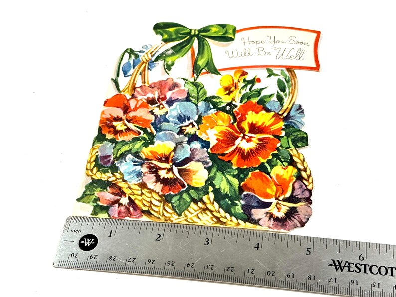 May include: A vintage greeting card featuring a basket overflowing with colorful pansies in shades of orange, yellow, blue, and purple. A green bow adorns the top, and a banner reads "Hope You Soon Will Be Well."