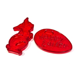 May include: Two red Easter-themed decorations. One is a bunny-shaped cookie cutter, and the other is an egg-shaped ornament with the words "Happy Easter" etched into it. Both are made of a translucent red material.