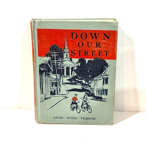 以下が含まれることがあります： 「Down Our Street」というタイトルのヴィンテージ本。赤と水色の表紙。表紙には、教会、家、2人の子供が自転車に乗っている通りのシーンのイラストが描かれています。著者の名前は一番下にあります。