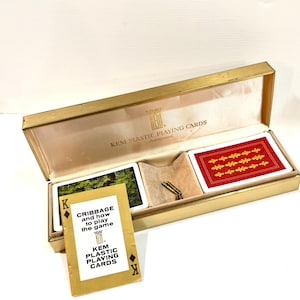 Pode incluir: Uma caixa retangular dourada contendo dois baralhos de cartas de plástico Kem, um com o verso vermelho estampado e outro com uma cena de floresta verde. Inclui dois pequenos pinos de metal e um livreto intitulado Cribbage and how to play the game.