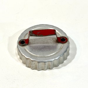 May include: A vintage, round cookie cutter with a fluted edge and a red handle. The metal cutter has a silver finish with some wear and tear, suggesting it's been used. The handle is a bright red color.