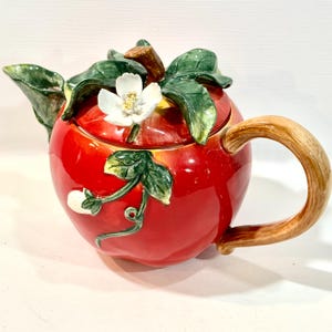 May include: A ceramic teapot shaped like a red apple, with green leaves and a white flower on top. The handle is a light brown color. The teapot is a decorative kitchen item.