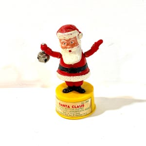 May include: A Santa Claus push button puppet featuring a red and white suit, a white beard, and a small silver bell held in one hand. The figure stands on a yellow cylindrical base with a label that reads SANTA CLAUS PUSH BUTTON PUPPET.