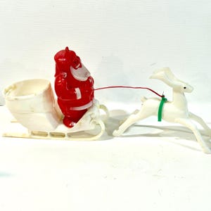 May include: A vintage plastic Christmas decoration featuring a red Santa Claus figure sitting in a white sleigh. A white plastic reindeer is attached to the sleigh by a thin red string and a green plastic harness, creating a festive holiday scene.