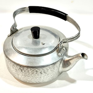 May include: A vintage aluminium tea kettle with a black handle and lid knob. The kettle has a textured body and a spout for pouring. The handle is arched and attached to the body with metal brackets. The kettle is designed for stovetop use.