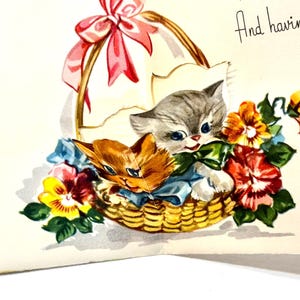 May include: A vintage greeting card illustration featuring two kittens nestled in a woven basket filled with colorful flowers. The basket is adorned with a pink bow. The text "And having lots" is written in cursive.