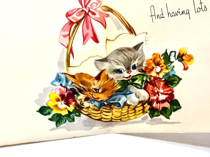 May include: A vintage greeting card illustration featuring two kittens nestled in a woven basket filled with colorful flowers. The basket is adorned with a pink bow. The text "And having lots" is written in cursive.