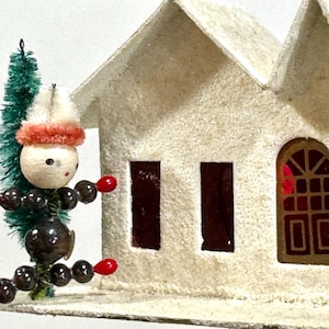Può includere: Un ornamento natalizio vintage con una casa bianca con superficie strutturata e una piccola figura. La figura ha una testa di legno, un corpo di perline nere e accenti rossi. Un piccolo albero verde è attaccato.