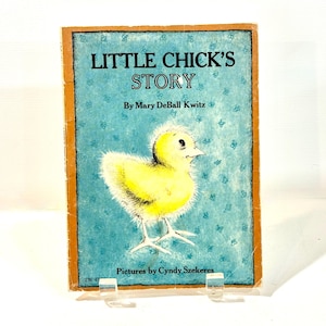 Puede incluir: Un libro infantil vintage titulado "Little Chick's Story" de Mary DeBall Kwitz, con ilustraciones de Cyndy Szekeres. La portada presenta un pollito amarillo sobre un fondo azul con patrones florales. El libro está sostenido por un soporte transparente.