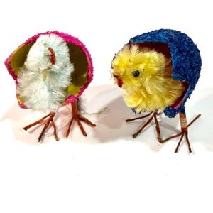 May include: Two vintage Easter decorations. One features a fluffy white chick in a pink and yellow eggshell, the other a yellow chick in a blue and red eggshell. Both have brown wire legs. A charming Easter theme.