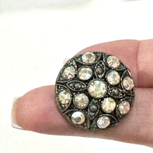 May include: A round, antique-style button with a silver-toned metal frame. The button features a pattern of clear, sparkling rhinestones in various sizes, arranged in a floral design. The button is held by a person's finger.