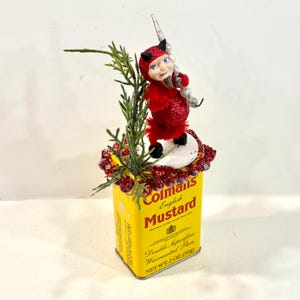 以下が含まれることがあります： 黄色いColman's English Mustardの缶の上に、小さな悪魔のフィギュアが乗った、気まぐれな手作りのアート作品。悪魔は赤色で、黒い角と銀色の三叉槍を持っています。缶には文字が書かれており、緑と赤いベリーで飾られています。