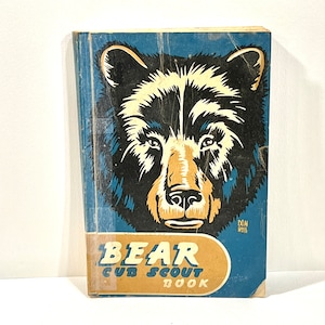 May include: A vintage Bear Cub Scout Book. The blue cover features a black and tan bear illustration. The title, "Bear Cub Scout Book," is in white and tan lettering. The book shows signs of wear and tear, indicating its age.