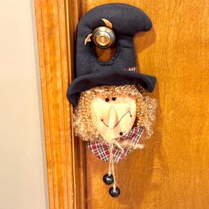 以下が含まれることがあります： 魔女の帽子を模した布製のドアハンガー。ぬいぐるみのような顔、カールしたブロンドの髪、チェック柄の襟が付いています。黒い帽子にはドアノブ用の穴があり、下部には細い紐で2つの小さな黒い鈴がぶら下がっています。