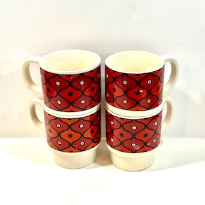 Könnte beinhalten: Vier gestapelte Keramikbecher mit einem rot-schwarzen geometrischen Muster. Die Becher haben weiße Henkel und einen cremefarbenen Sockel. Das Muster zeigt schwarze Ovale mit weißen Punkten auf rotem Grund.