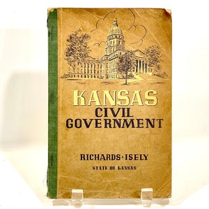 May include: A vintage hardcover book titled Kansas Civil Government by Richards and Isely. The tan cover features a black line drawing of the Kansas State Capitol building, with green fabric binding on the spine and black text throughout.