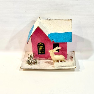 Puede incluir: Una pequeña casa rosa con un tejado blanco y azul, una ventana negra y una figura de ciervo. La casa tiene un pequeño arbusto y se asienta sobre una base blanca, creando una escena invernal.