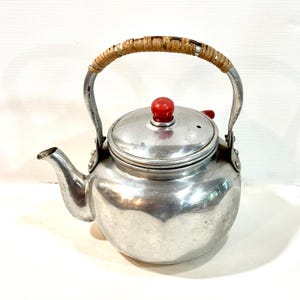 Peut inclure: Une bouilloire à thé vintage en aluminium avec une poignée tressée et un bouton rouge sur le couvercle. La bouilloire a un bec verseur et est conçue pour une utilisation sur la cuisinière. La poignée est en métal et en matériau tressé.