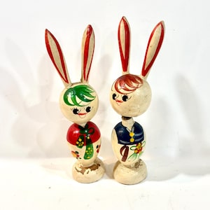 May include: Two hand-painted wooden rabbit figurines with tall ears. The left rabbit has green hair and a red outfit with yellow accents. The right rabbit has brown hair and a blue outfit with yellow buttons. Both have cream-colored bases.
