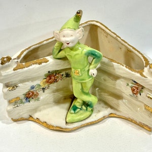 May include: A vintage ceramic planter with a whimsical elf figurine in a green outfit. The planter has a cream base with gold trim and floral accents. The elf leans against the planter, adding a playful touch to this decorative piece.