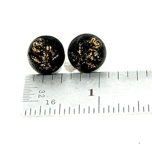 May include: Two round black buttons featuring a raised gold-colored floral design on the surface. The buttons are positioned next to a metal ruler, showing each button has a diameter of approximately 0.5 inches.