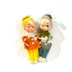Peut inclure: Deux décorations de Noël vintage en forme d'elfes. L'un porte une veste orange avec des boutons blancs, un pantalon jaune et un chapeau jaune. L'autre porte un chapeau gris, un col vert, une salopette grise et une chemise blanche. Les deux ont les cheveux blonds et les joues roses.