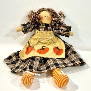 May include: A handmade doll with a fabric body, wearing a plaid dress and an apron with pumpkin designs. The doll has curly brown hair and orange and white striped legs. A charming fall-themed decorative item.