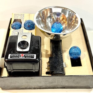 May include: A vintage Brownie Hawkeye camera flash model kit. The kit includes a black camera, a chrome flash reflector, and several blue glass flash bulbs. The camera and flash are in a cardboard box.
