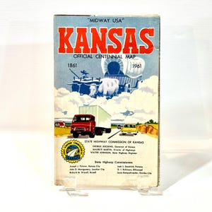 Puede incluir: Un mapa oficial del centenario de Kansas de 1961. La portada presenta un título rojo y blanco, ilustraciones de una carreta cubierta, ganado y un camión rojo. El mapa se muestra en un soporte transparente.