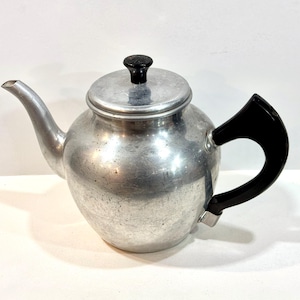 May include: A vintage aluminum teapot with a black handle and lid knob. The teapot has a rounded body, a curved spout, and a lid. The handle is a curved, solid piece. The teapot is sitting on a white surface.