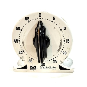 Può includere: Timer da cucina bianco vintage con dettagli neri. Il timer presenta un quadrante in bianco e nero con numeri da 0 a 55 e una manopola nera. La base del timer riporta la scritta "Mark-Eric Corporation".