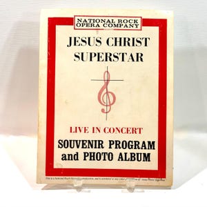 Könnte beinhalten: Ein rechteckiges Souvenirprogramm und Fotoalbum für Jesus Christ Superstar Live in Concert von der National Rock Opera Company. Das Cover ist cremefarben mit einem roten Rand, schwarzem Text und einem rosa Violinschlüssel mit Kreuz.
