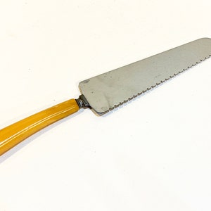 May include: A vintage cake server. It has a stainless steel blade with a serrated edge. The handle is a light amber colour. The server is designed for cutting and serving cakes and pastries.