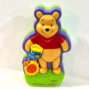 May include: A yellow Winnie the Pooh electronic toy wearing a red shirt, set against a purple background. Pooh holds a blue honey pot labeled FIND IDENTIFY OFF and stands next to a yellow sunflower speaker. The base reads Pooh Smart Sticks.