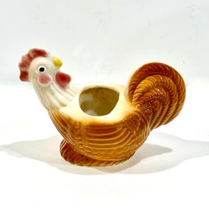 May include: A ceramic rooster-shaped planter with a cream-colored head and a brown body. The rooster has a red comb and wattle, a yellow beak, and a round opening on its back for planting. The planter is a decorative item.