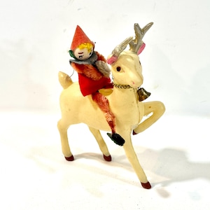 Op de afbeelding: Een vintage kerstdecoratie met een crèmekleurig rendier met gouden geweien en een kleine belhalsband. Een kleine elf in een rode outfit en hoed rijdt op de rug van het rendier. De elf heeft een blond hoofd en een grijze bontkraag.